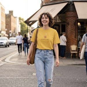 Puede incluir: Una camiseta de manga corta color amarillo mostaza con un pequeño gráfico de sol rojo y el texto "you're about day right.", combinada con vaqueros azul claro desgastados y un bolso de lona marrón al hombro.