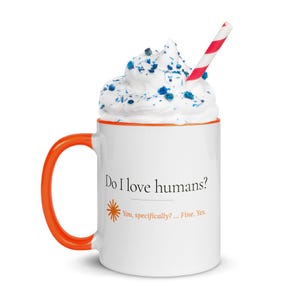 May include: A white ceramic mug with an orange handle and rim, featuring the black text "Do I love humans?" and orange text "You, specifically? ... Fine. Yes." with a star icon. The mug is filled with white whipped cream, blue sprinkles, and a red and white striped straw.