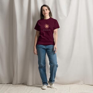 May include: A burgundy short-sleeved t-shirt featuring a light pink sunburst graphic on the chest with the text "you're absolutely right" printed underneath in small white letters, paired with blue denim jeans and white sneakers.
