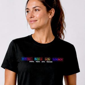 May include: A black short-sleeved t-shirt featuring a graphic design with colorful square icons containing letters that spell out "Di Gi Ta L Me Di A Ar Ts Te Ac He R" and the text "DIGITAL MEDIA ARTS TEACHER" printed in white underneath the icons.