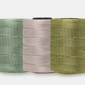 May include: Three cylindrical spools of cord arranged side by side. The left spool is sage green, the center spool is a pale dusty pink, and the right spool is olive green. Each spool features a tightly wound, smooth, glossy synthetic cord.