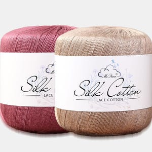 Pode incluir: Dois novelos de fio cilíndricos, um em rosa escuro e outro em bege claro, cada um envolto numa faixa de papel branco com o texto Silk Cotton LACE COTTON e um gráfico de nuvem com ilustrações florais.