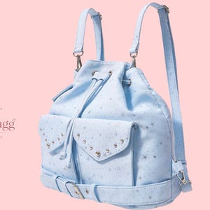 Op de afbeelding: Een lichtblauwe rugzak met trekkoord, een zilveren sterrenpatroon, twee voorzakken met zilveren studs, verstelbare schouderbanden en een gespriem aan de onderkant. Het logo LioraBagg Handmade Bags is zichtbaar aan de linkerkant.
