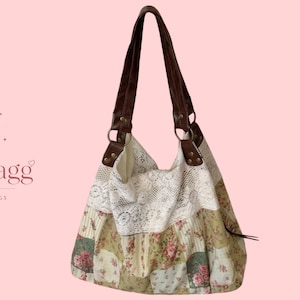 Puede incluir: Un bolso de hombro estilo hobo con un cuerpo de tela de patchwork floral en tonos verde, crema y rosa. El bolso presenta un borde ancho de encaje blanco en la parte superior y dos correas de piel sintética marrón oscuro con anillas.