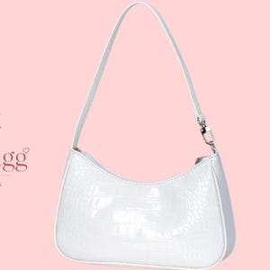 May include: A white shoulder bag with a glossy, textured crocodile-embossed finish. The bag features a curved top silhouette, a single thin strap attached with silver-toned metal hardware, and a zipper closure. The text LioraBagg Handmade Bags is visible.