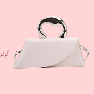 Peut inclure: Un sac à main blanc avec une forme trapézoïdale distinctive et un rabat avant incurvé. Le sac présente une poignée métallique épaisse et polie de couleur argentée, ainsi qu'une bandoulière amovible. Le logo LioraBagg Handmade Bags est visible.