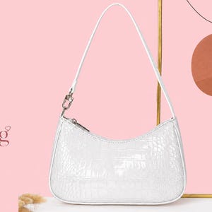 May include: A white shoulder bag with a glossy crocodile skin texture, featuring a single thin strap attached with silver-toned metal hardware and a top zipper closure. The bag has a curved, crescent-shaped silhouette and a smooth white trim.