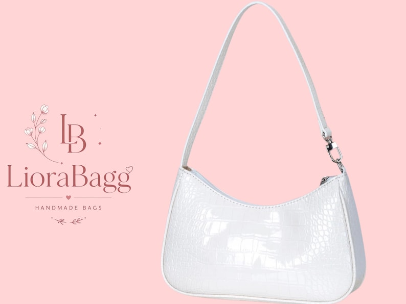 May include: A white shoulder bag with a glossy, textured crocodile-embossed finish. The bag features a curved top silhouette, a single thin strap attached with silver-toned metal hardware, and a zipper closure. The text LioraBagg Handmade Bags is visible.