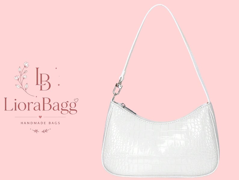 May include: A white shoulder bag with a textured crocodile-embossed pattern, featuring a single thin strap, a curved top zipper closure, and silver-toned hardware. To the left, the text reads LB LioraBagg HANDMADE BAGS with floral accents.