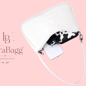 May include: A white shoulder bag with a crocodile-embossed texture and a single strap. The bag is open, revealing a white smartphone, a black and white cow-print wallet, and a black object inside. The text LioraBagg HANDMADE BAGS is visible.