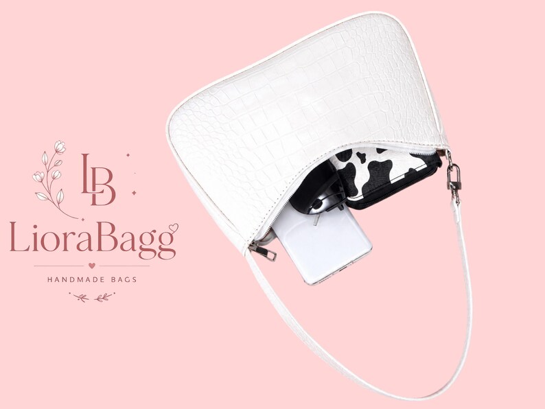 May include: A white shoulder bag with a crocodile-embossed texture and a single strap. The bag is open, revealing a white smartphone, a black and white cow-print wallet, and a black object inside. The text LioraBagg HANDMADE BAGS is visible.