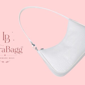 May include: A white shoulder bag with a textured crocodile skin pattern, a single strap, and a silver-toned zipper closure. The text LioraBagg HANDMADE BAGS with a floral logo is visible to the left of the bag on a light pink background.