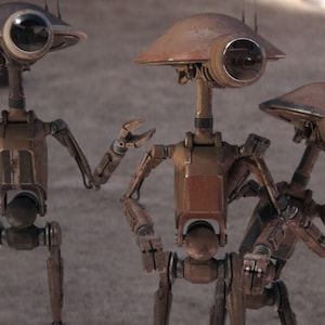 May include: Three rust-colored robotic figures with dome-shaped heads and single large circular optical sensors. Each droid features a slender neck, articulated arms with multi-fingered hands, and a central torso structure with visible mechanical joints.