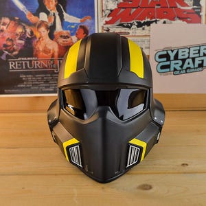 May include: A matte black full-face helmet featuring a dark tinted visor and prominent yellow racing stripes running from the crown to the sides. The lower section includes silver-colored vent grilles framed by yellow accents on both sides.