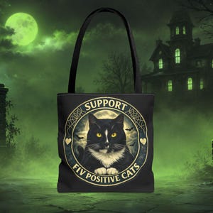 Może przedstawiać: Czarna torba typu tote z okrągłą grafiką przedstawiającą czarno-białego kota z żółtymi oczami. Napis głosi SUPPORT FIV POSITIVE CATS. Wzór zawiera pajęczyny, nietoperze oraz pełnię księżyca w tle wewnątrz okrągłej ramki.