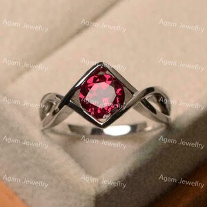 May include: A silver-toned ring featuring a round, faceted, deep red gemstone set within a diamond-shaped frame. The band consists of two intertwining, polished metal strands that meet at the central setting, creating a continuous, fluid design.