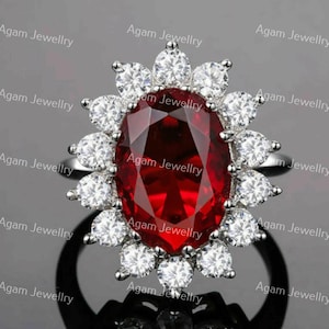 May include: A silver-toned ring featuring a large, oval-cut deep red gemstone at the center, surrounded by a halo of fourteen round, clear white stones. The text Agam Jewellry is repeated multiple times across the image as a watermark.