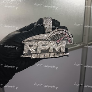 May include: A silver-toned pendant featuring the text RPM and DIESEL encrusted with clear stones. The design includes a tachometer gauge graphic with black stones and a red needle accent. The bail is also covered in clear stones.