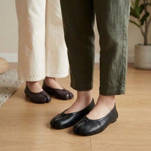 Könnte beinhalten: Zwei Paar Ballerinas mit geteilter Zehenpartie aus glattem Leder. Ein Paar ist einfarbig schwarz und das andere Paar ist dunkelbraun. Jeder Schuh hat eine abgerundete Silhouette mit einer markanten vertikalen Naht in der Mitte.