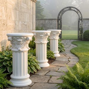 May include: Three white cylindrical pedestals featuring fluted columns and ornate capitals with carved leaf and berry motifs. Each pedestal has a circular top surface and a wide, stable base, arranged in a row along a stone garden pathway.