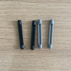 May include: Four elongated, cylindrical silicone temple tips for eyeglasses, arranged vertically. Two of the tips are solid black, and two are light gray. Each piece features a narrow, centered rectangular slot running along the length of the body.
