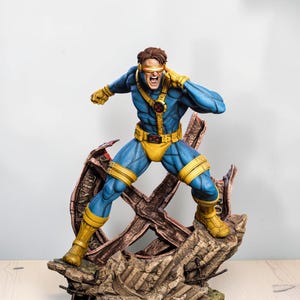 May include: A detailed figurine of a character in a blue and yellow suit with a red visor, standing in a dynamic pose on a rocky, debris-filled base. The character has brown hair, a yellow belt with a red buckle, and is touching the side of the visor.