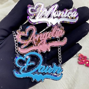 May include: Three custom name necklaces featuring layered acrylic designs. The top piece is silver-toned with the name Monica, the middle is light pink with the name Angelia, and the bottom is light blue with the name Dawn. Each includes heart accents.