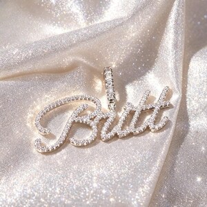 May include: A silver-toned pendant featuring the name Britt written in a cursive script font. The entire surface of the letters and the vertical bail at the top are encrusted with numerous small, clear, sparkling stones that reflect light.