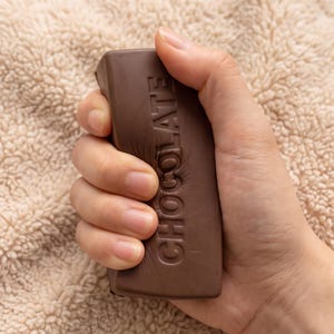 May include: A rectangular, dark brown, flexible bar with the word CHOCOLATE embossed vertically in capital letters. The material appears soft and pliable, showing visible indentations and texture changes where it is being firmly gripped.