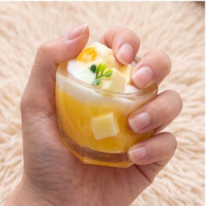 May include: A small, hexagonal glass container filled with a yellow, translucent gel substance. The top layer features white, opaque, cube-shaped inserts, a small green plastic leaf decoration, and a single orange, circular fruit-like accent piece.