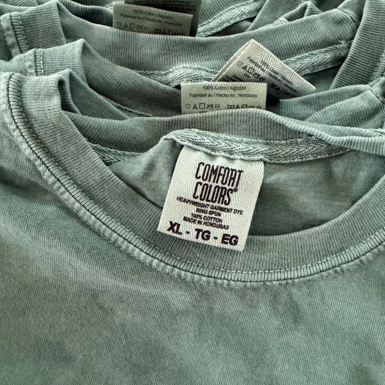 May include: A stack of sage green t-shirts featuring a white interior neck label with the text Comfort Colors, Heavyweight Garment Dye, Ring Spun, 100% Cotton, Made in Honduras, and the size XL-TG-EG printed in black ink on the fabric tag.