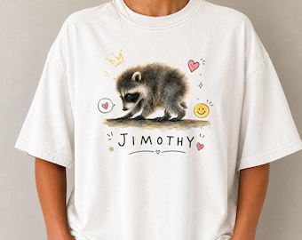 May include: A white short-sleeved t-shirt featuring a central graphic of a grey raccoon. The design includes the name JIMOTHY in black capital letters, a yellow crown, a yellow smiley face, and several small pink hearts and yellow stars.