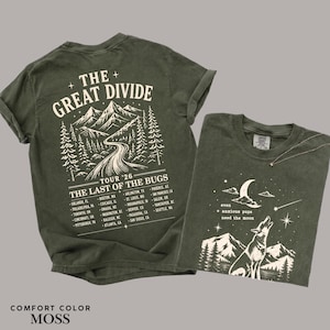 May include: An olive green t-shirt featuring a back graphic of mountains, a winding road, and the text THE GREAT DIVIDE TOUR '26 THE LAST OF THE BUGS with a list of cities. The front graphic shows a wolf howling at a crescent moon and mountains.