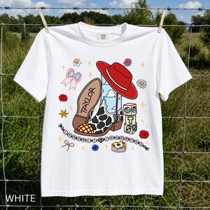 May include: A white short-sleeve t-shirt featuring a graphic print of a cow-print cowboy boot, a red cowboy hat, blocks with the letters T, S, and a heart, and a beaded string with the text I KNEW IT I KNEW YOU and the name TAYLOR.