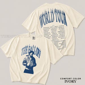 May include: An ivory short-sleeve t-shirt featuring a blue graphic print. The front displays a person in a hooded jacket with the text THE FALL OFF. The back shows the text WORLD TOUR 2024 followed by a list of cities and locations in blue.