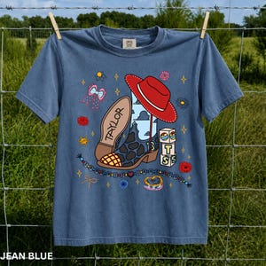 May include: A blue short-sleeve t-shirt featuring a graphic print of a cowboy boot with the name Taylor, a red cowboy hat, blocks with the letters T S, a beaded string with the text I KNEW IT I KNEW YOU, and various small floral and star icons.