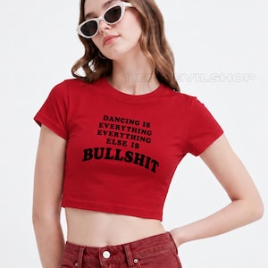 May include: A red short-sleeved crop top featuring black text that reads DANCING IS EVERYTHING EVERYTHING ELSE IS BULLSHIT. The garment has a crew neckline and a fitted silhouette, paired with white cat-eye sunglasses and red denim bottoms.