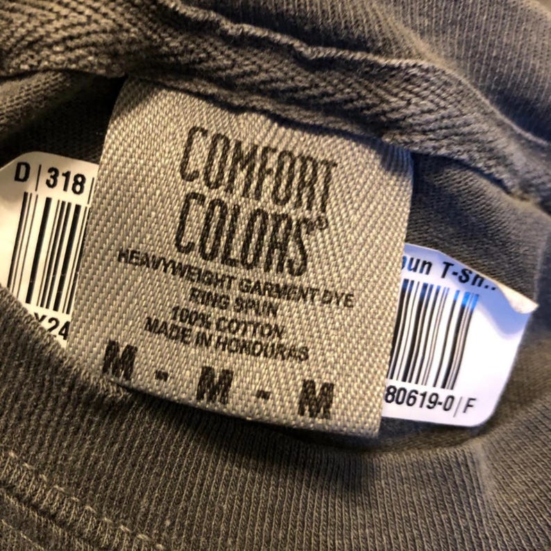 May include: A gray garment tag with black text reading COMFORT COLORS, HEAVYWEIGHT GARMENT DYE, RING SPUN, 100% COTTON, MADE IN HONDURAS, and size M. The tag is attached to a dark gray fabric with two white barcode stickers on either side.