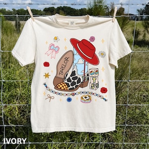 May include: An ivory t-shirt featuring a graphic print of a cow-print cowboy boot, a red hat, and blocks spelling BTS. The design includes the name Taylor, a beaded string with the text I KNEW IT I KNEW YOU, a pink bow, and flowers.