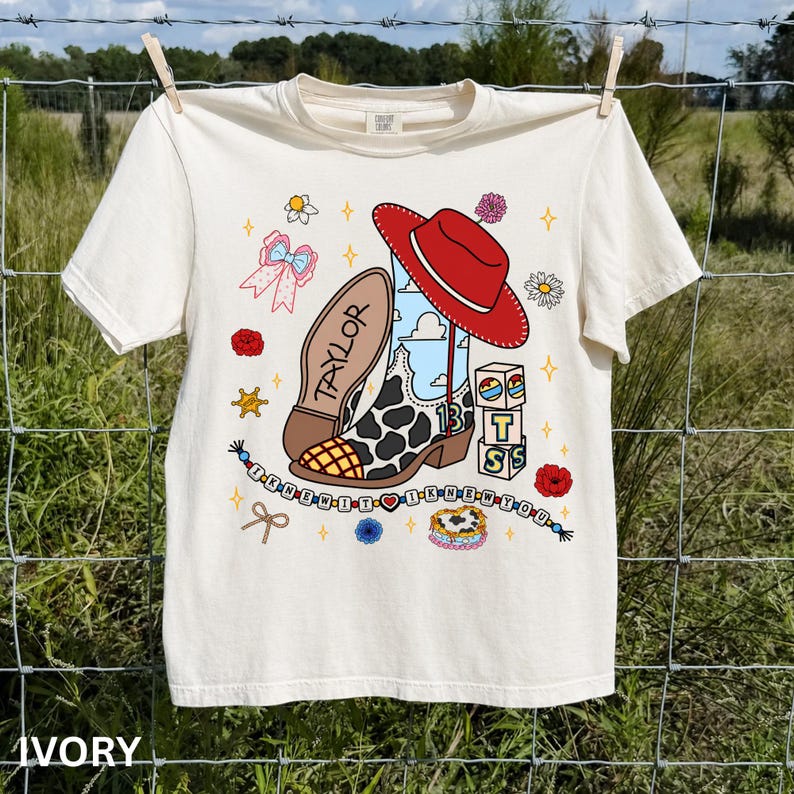 May include: An ivory t-shirt featuring a graphic print of a cow-print cowboy boot, a red hat, and blocks spelling BTS. The design includes the name Taylor, a beaded string with the text I KNEW IT I KNEW YOU, a pink bow, and flowers.