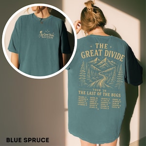 May include: A blue spruce t-shirt featuring a small front graphic and a large back graphic with the text The Great Divide, The Last of the Bugs, Tour '26, and a list of cities. The back design includes a mountain range, pine trees, and a road.