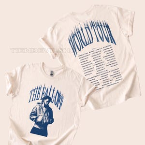 May include: A cream-coloured t-shirt featuring a blue graphic of a person in a jacket with the text THE FALL OFF. The back of the shirt displays the text WORLD TOUR in blue above a list of cities and the year 2024 in a matching blue font.
