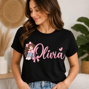 May include: A black short-sleeved t-shirt featuring a graphic of a cartoon girl with brown hair, a pink bow, and a pink sweater. The text "Olivia" is printed in large pink script lettering next to the character, accented by a pink bow and heart.