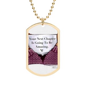 Pode incluir: Um pingente de placa de identificação dourado em uma corrente de bolinhas correspondente. O pingente apresenta uma ilustração de uma pessoa com um suéter de tricô roxo segurando uma caneca branca com o texto Your Next Chapter Is Going To Be Amazing e um coração.