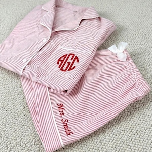 Pode incluir: Um conjunto de pijama às riscas cor-de-rosa e brancas composto por uma camisa de botões com colarinho e calções. A camisa apresenta um bolso no peito bordado com o monograma vermelho AGC, enquanto os calções têm o texto vermelho Mrs. Smith.