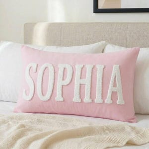 Könnte beinhalten: Ein rechteckiges, hellrosa Dekokissen mit dem Namen SOPHIA in großen, weißen, strukturierten Chenille-Buchstaben im Varsity-Stil, die mittig auf der Vorderseite angebracht sind. Das Kissen hat eine glatte Stoffoberfläche und liegt horizontal auf einem Bett.