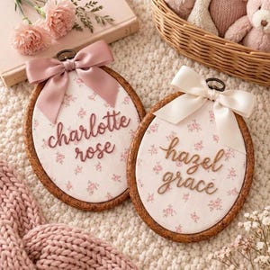 Puede incluir: Dos bastidores de bordado ovalados de madera con tela blanca de estampado floral rosa. Un bastidor muestra el nombre charlotte rose en letra marrón con un lazo de satén rosa empolvado, y el otro muestra hazel grace con un lazo de satén crema.