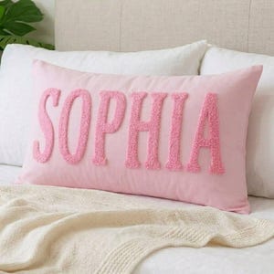 Könnte beinhalten: Ein rechteckiges, hellrosa Dekokissen mit dem Namen SOPHIA in großen, strukturierten, erhabenen Buchstaben im Chenille-Stil in der Mitte. Das Kissen hat eine glatte Stoffoberfläche und liegt horizontal auf einem Bett mit weißer Bettwäsche.
