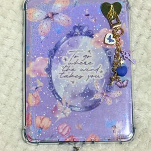 May include: A rectangular purple tablet case with a glittery finish, featuring a central ornate mirror frame graphic with the text To go where the wind takes you. The case is decorated with butterflies, clouds, and fairies, and includes a gold-toned charm chain with a heart, a blue butterfly, a purple key, a blue heart, a moon, and a purple butterfly.