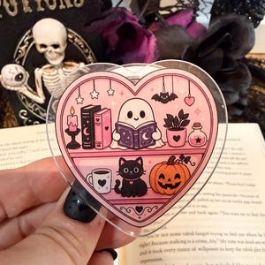 May include: A heart-shaped acrylic charm featuring a pink illustration of a ghost reading a book on a shelf with a candle, books, a plant, and a potion bottle. The lower shelf displays a mug, a black cat, and a jack-o'-lantern pumpkin.